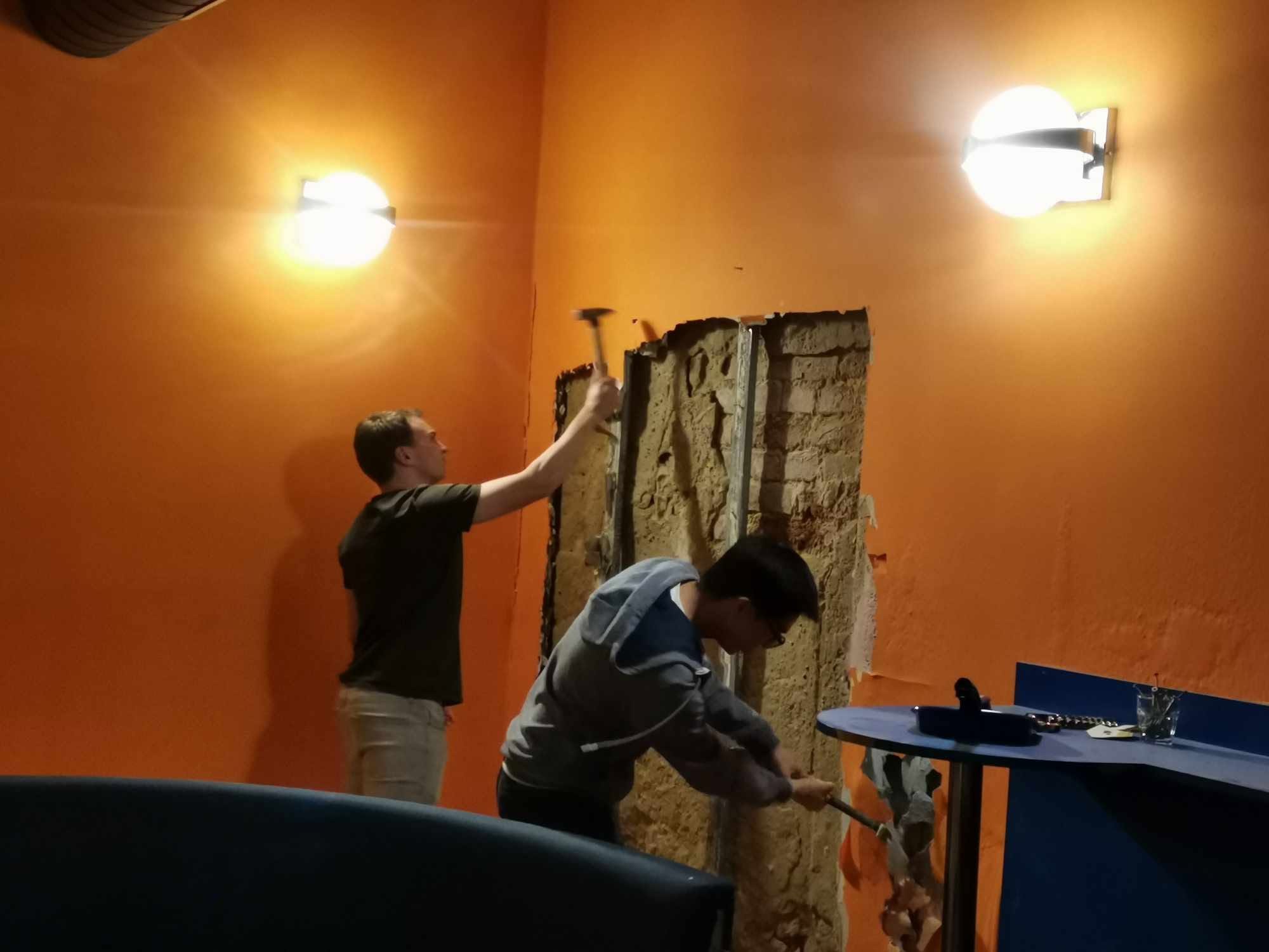 Two people chipping plaster off the orange wall